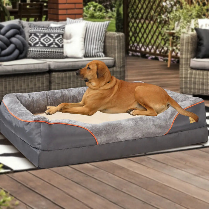 A dog resting on an orthopedic foam dog bed with a gray cover and orange detailing. The bed is placed on a wooden deck, and there is outdoor furniture in the background.